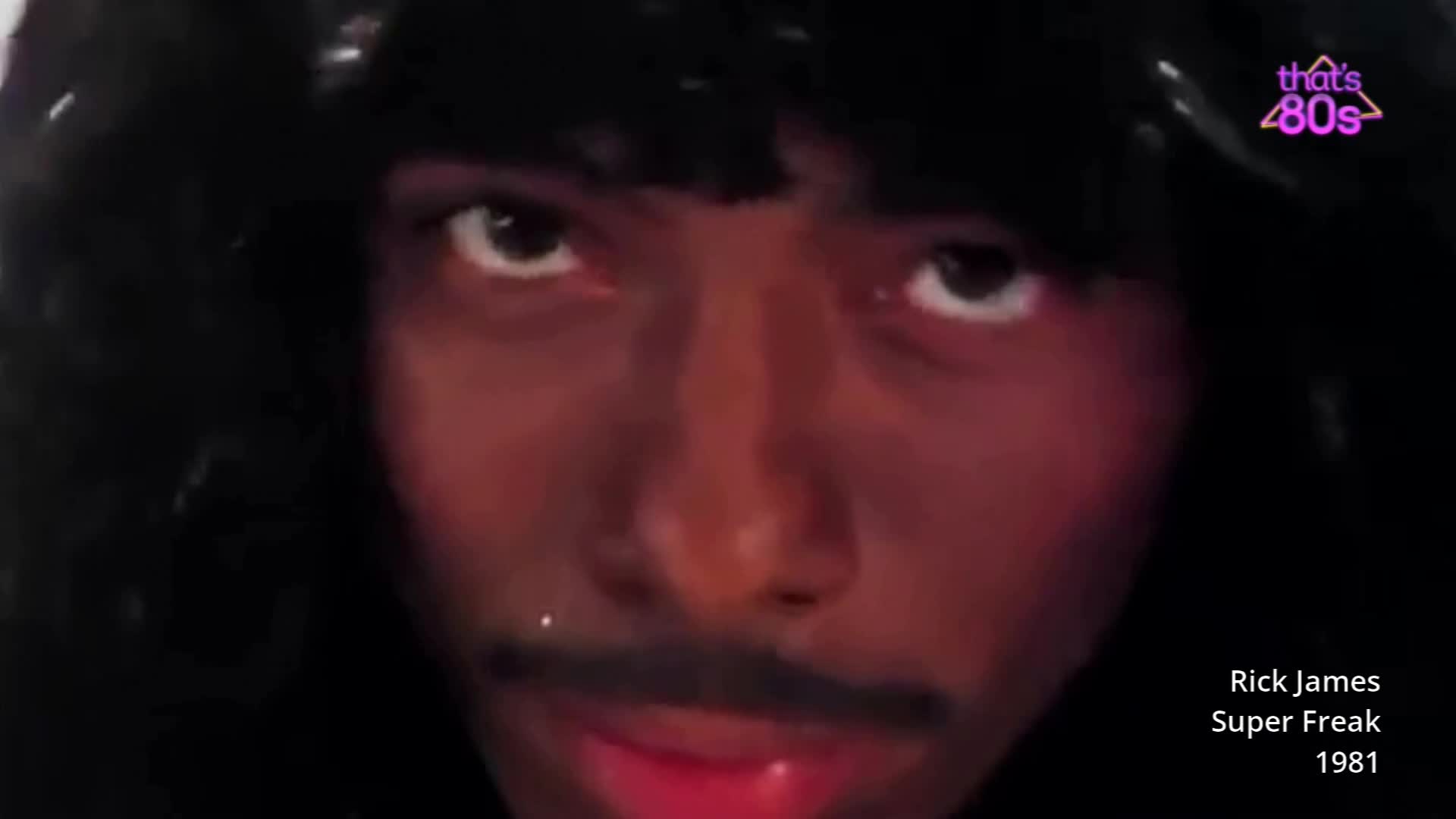 A man with a dark, voluminous wig and a thin moustache stares directly into the camera. His eyes are wide and intense, framed by heavy eyeliner, and his lips are a glossy, vibrant red. A man with a dark, voluminous wig and a thin moustache stares directly into the camera. His eyes are wide and intense, framed by heavy eyeliner, and his lips are a glossy, vibrant red.