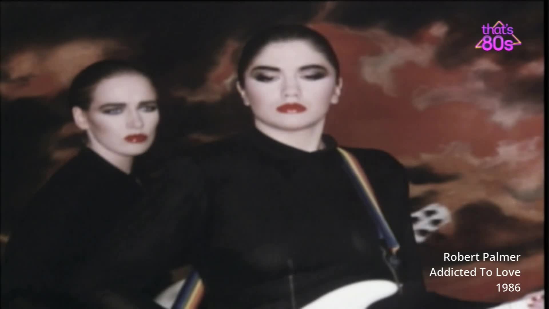 Two women with dramatic makeup and black outfits stand with guitars. The one in front stares ahead, while the one behind looks to the side with a stern expression. Two women with dramatic makeup and black outfits stand with guitars. The one in front stares ahead, while the one behind looks to the side with a stern expression.