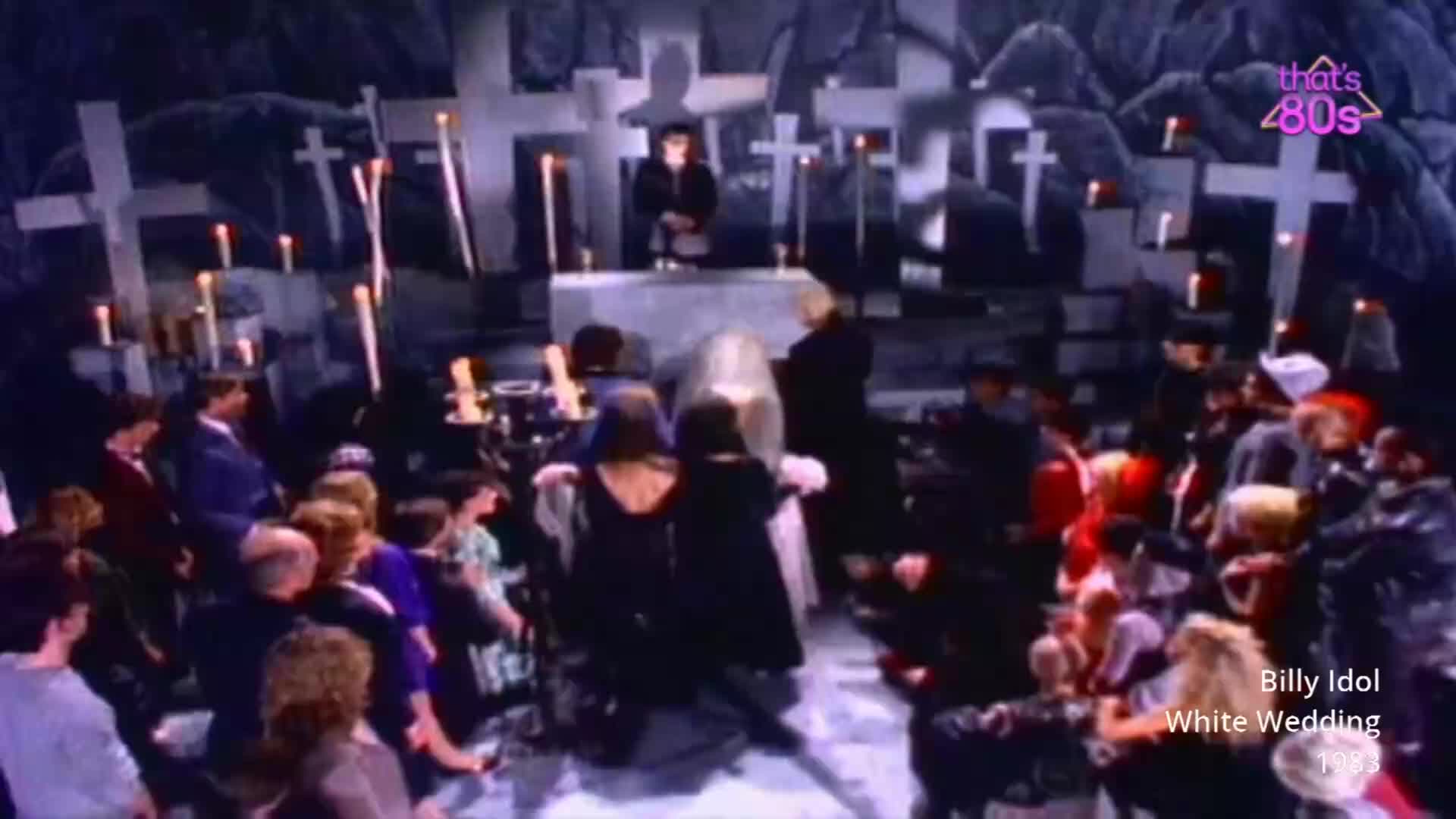A figure in black stands before a stone altar, surrounded by a crowd gathered amongst stark white crosses. Candles flicker, casting a dim light on the scene. A figure in black stands before a stone altar, surrounded by a crowd gathered amongst stark white crosses. Candles flicker, casting a dim light on the scene.