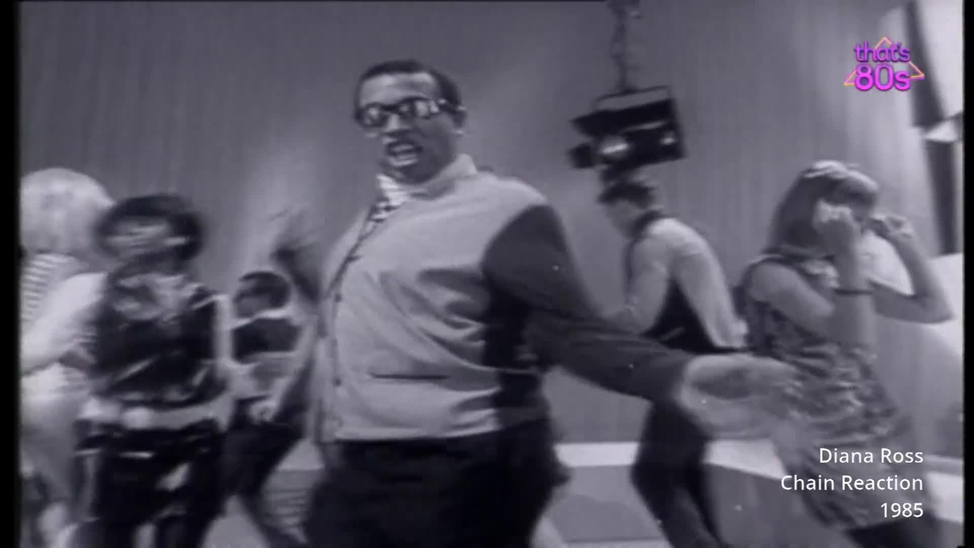 A man in a suit and glasses is dancing energetically, his arms outstretched. Behind him, other people are moving to the music on a television studio floor.