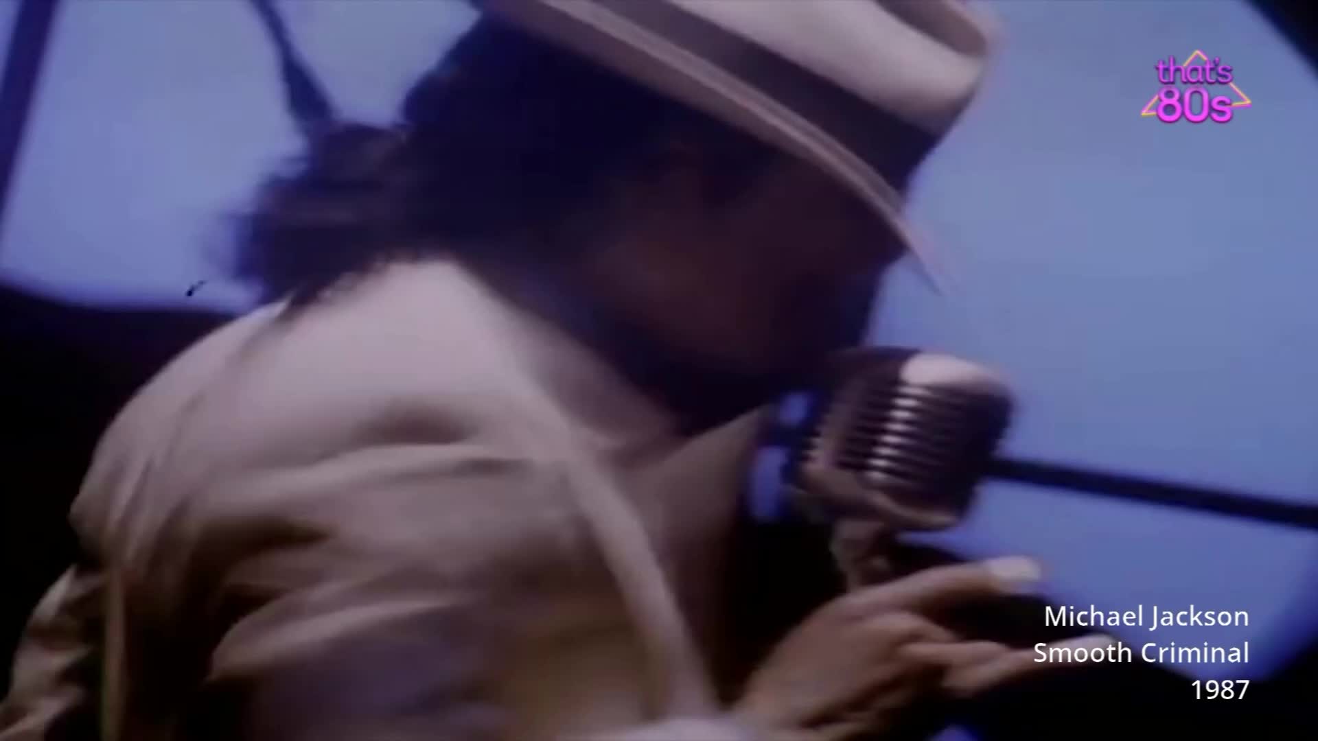 Michael Jackson leans into a vintage microphone, his fedora tilted low. His light-coloured suit jacket is a sharp contrast to the deep blue background.