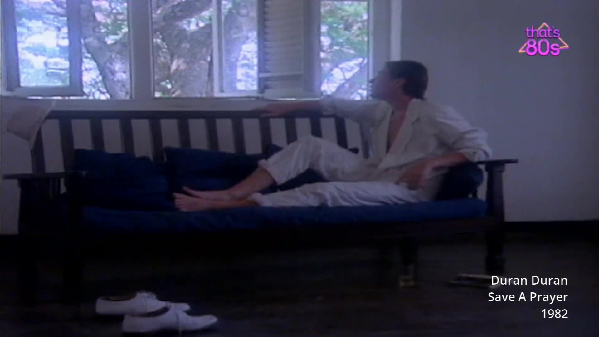 A man in a white jumpsuit reclines on a dark blue sofa by a window, his arm resting on the backrest. A pair of white tennis shoes sits on the floor in front of the sofa.