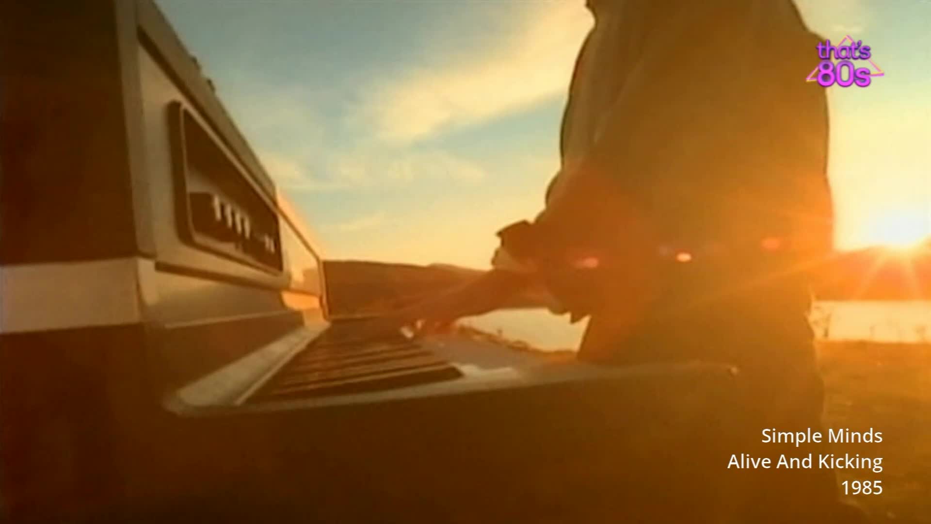 A man plays a keyboard outdoors as the sun sets over a body of water. The warm light catches the keys and his hands as he presses them.