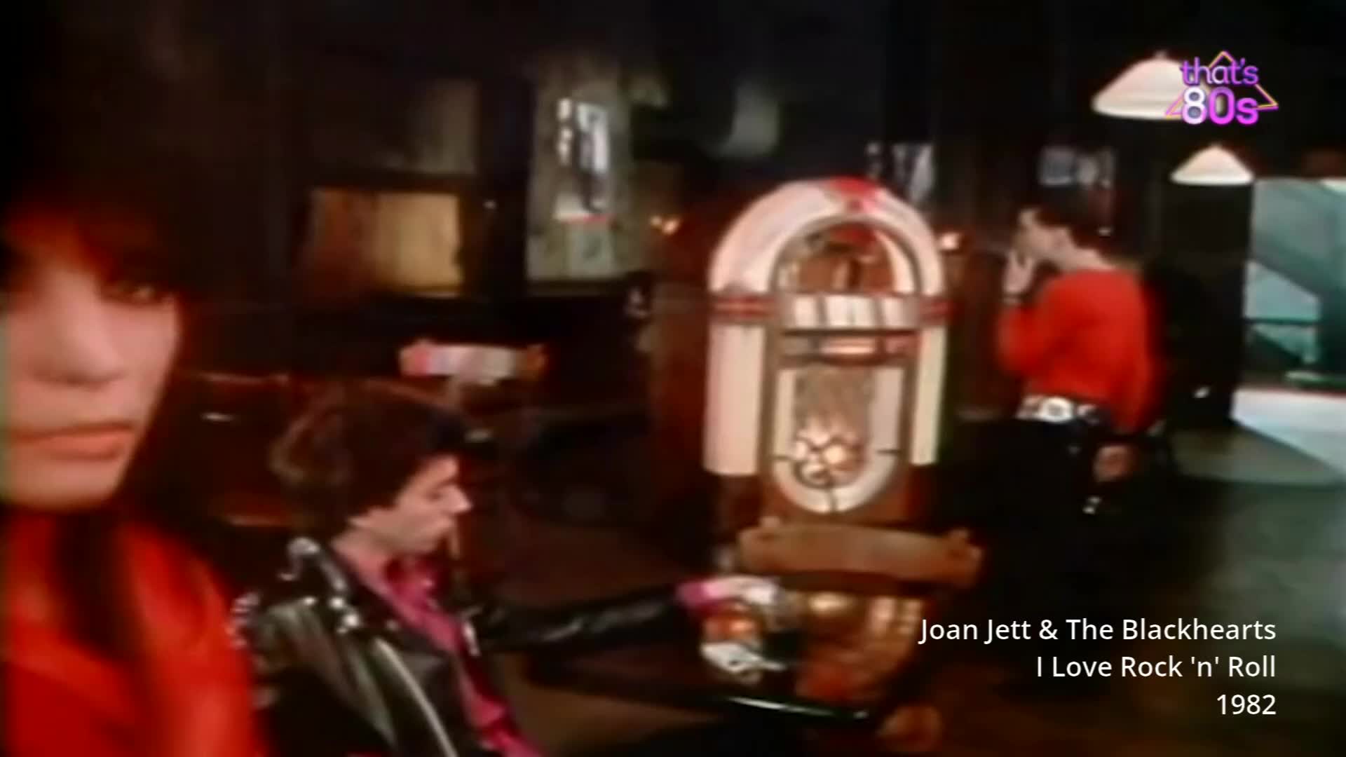 A woman with dark hair leans forward, her red top a stark contrast to the dim interior. Beside her, a man in a black leather jacket looks away, his profile sharp against the glow of a vintage jukebox.