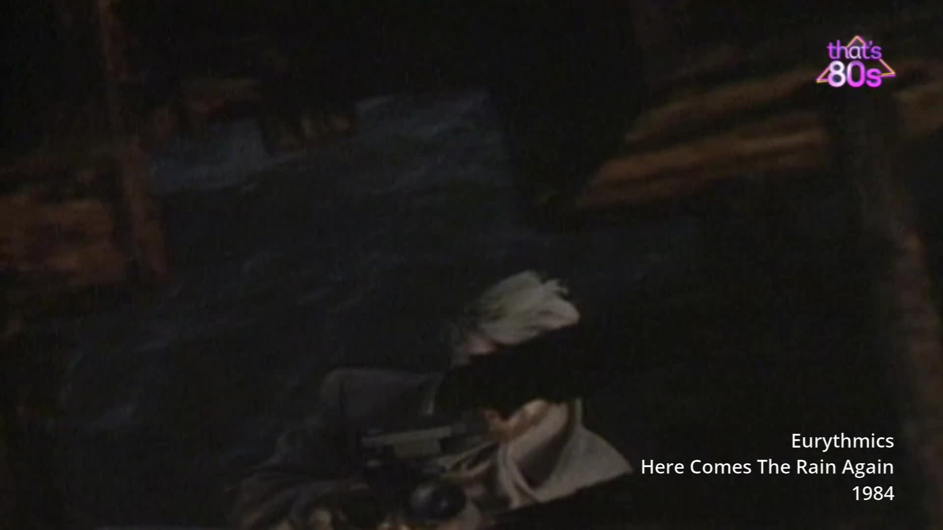 A person with light-coloured hair is looking into a camera, their face partially obscured by shadow. The scene is dark and moody, with a hint of the 1980s aesthetic.