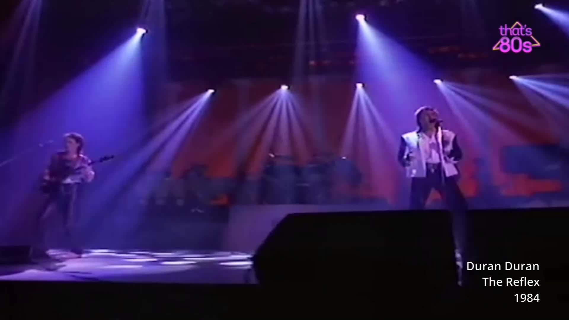 A guitarist stands bathed in blue light, his instrument slung low. The lead singer, in a sharp white jacket, belts into a microphone under a dramatic spotlight.