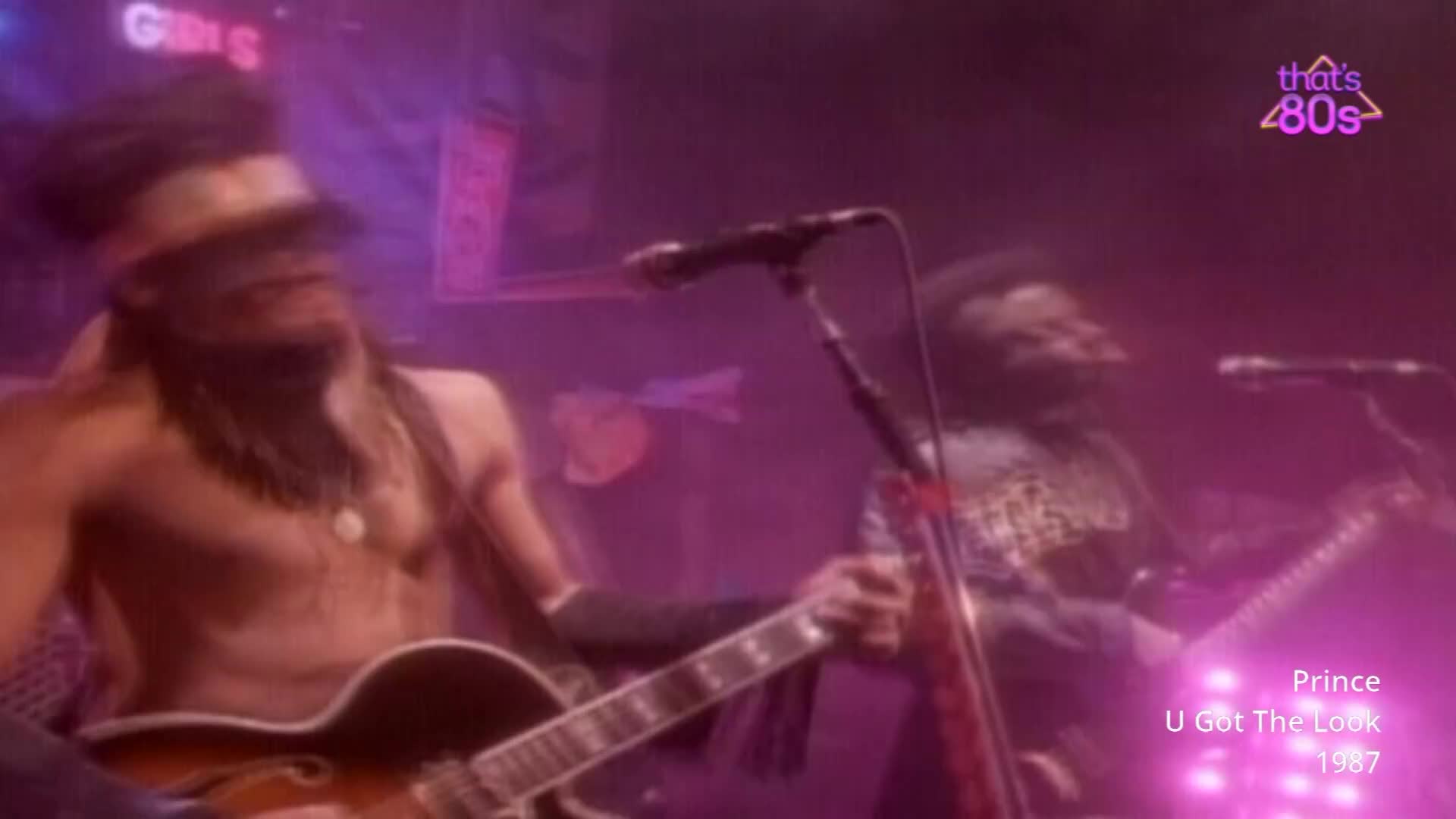 A guitarist with a headband and beads is playing a hollow-body electric guitar. Another musician, also with a guitar, stands to his right.