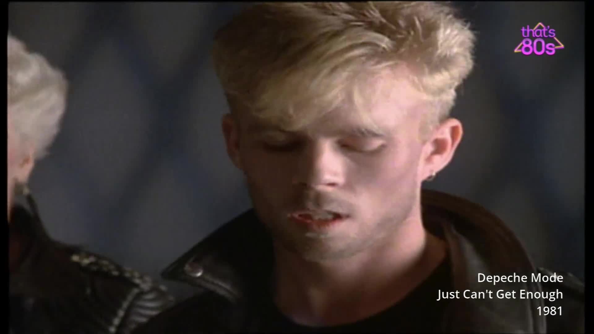 A young man with bleached blonde hair stands with his eyes closed, his mouth slightly open. He wears a black leather jacket, a common sight on the UK music scene of the early 1980s.