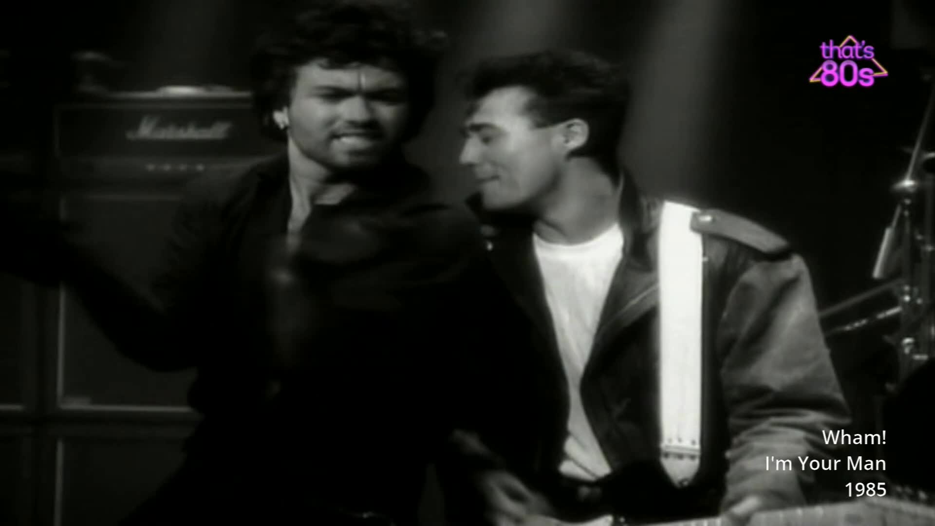 Two blokes are on stage, one with dark, curly hair is singing into a microphone. The other, with slicked-back hair, is holding a guitar strap.