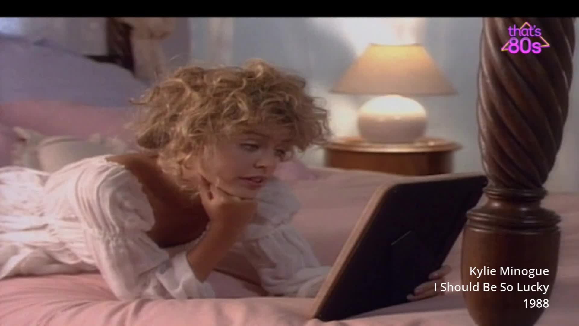 A young woman with curly blonde hair lies on a pink bed, holding a framed photograph. She's wearing a white, frilly nightgown, typical of the late 80s.