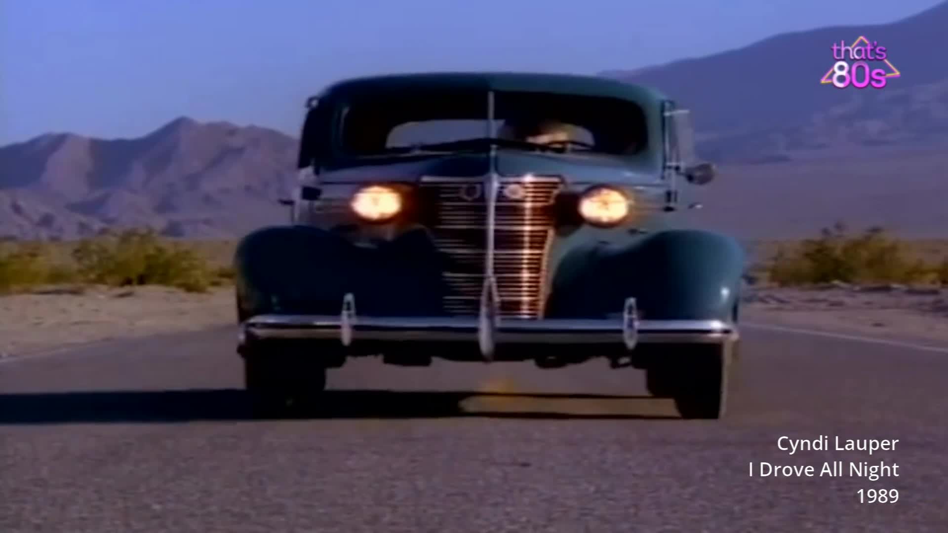 A dark green vintage car drives towards me on a deserted road. Its headlights cut through the twilight, illuminating the dusty desert landscape.
