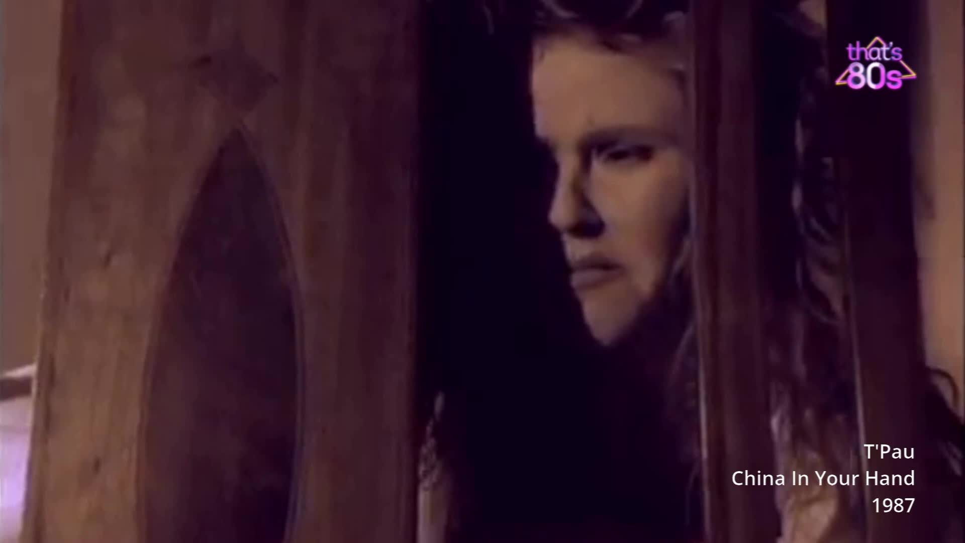 A young woman with blonde hair stares intently from behind a wooden frame. The lighting is dim, casting long shadows across her face, and the "That's 80s" logo is visible in the corner.