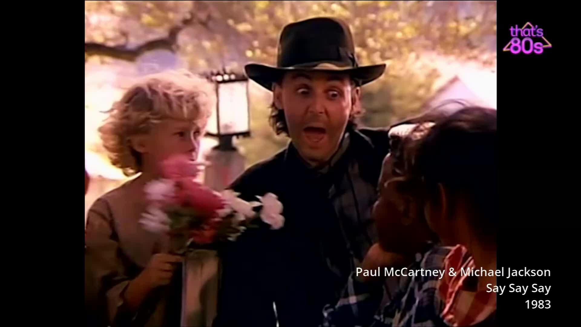 A man in a black hat stares with wide eyes, his mouth open in surprise. A young girl with curly blonde hair holds a bouquet of pink and white flowers.