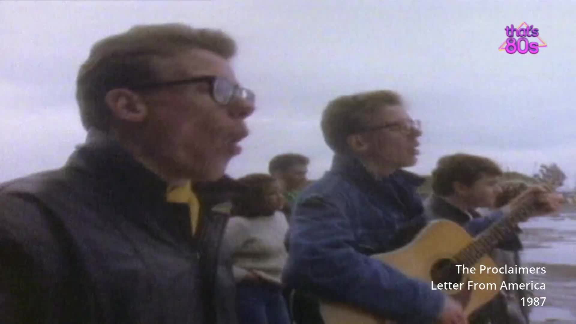Two men in glasses are singing, one with an acoustic guitar. They stand on a beach with a grey sky overhead.