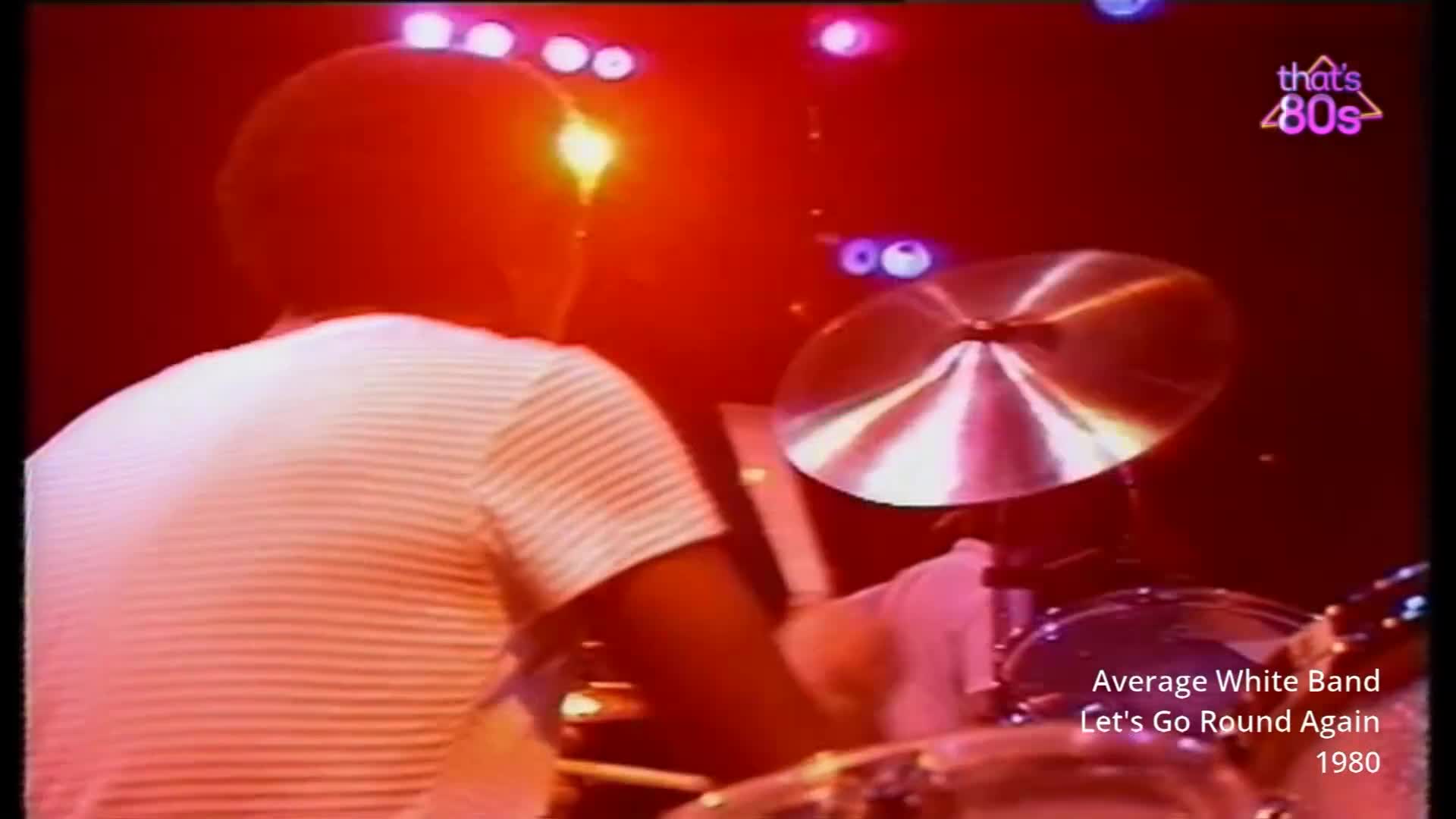 The drummer's white-striped shirt is illuminated by the stage lights as he hits the cymbal. The polished metal of the cymbal catches the vibrant red and purple hues of the lighting rig above.