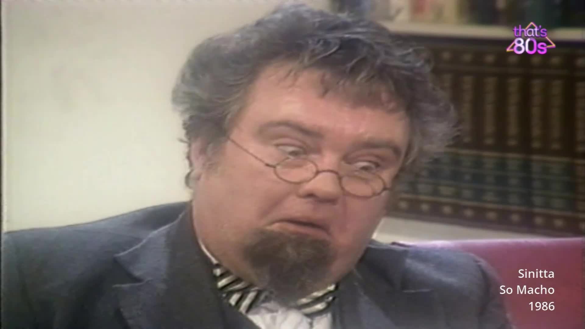 A man with a surprised expression and round glasses leans forward. His dark suit jacket and striped bow tie suggest a formal setting from the mid-1980s.