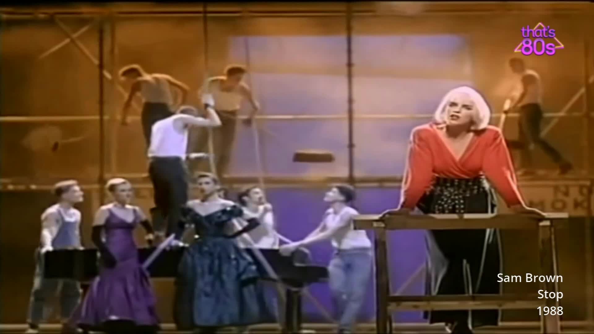 A woman with bright blonde hair, wearing a red top and a studded skirt, leans on a wooden table. Behind her, several men in tank tops and trousers move around scaffolding.