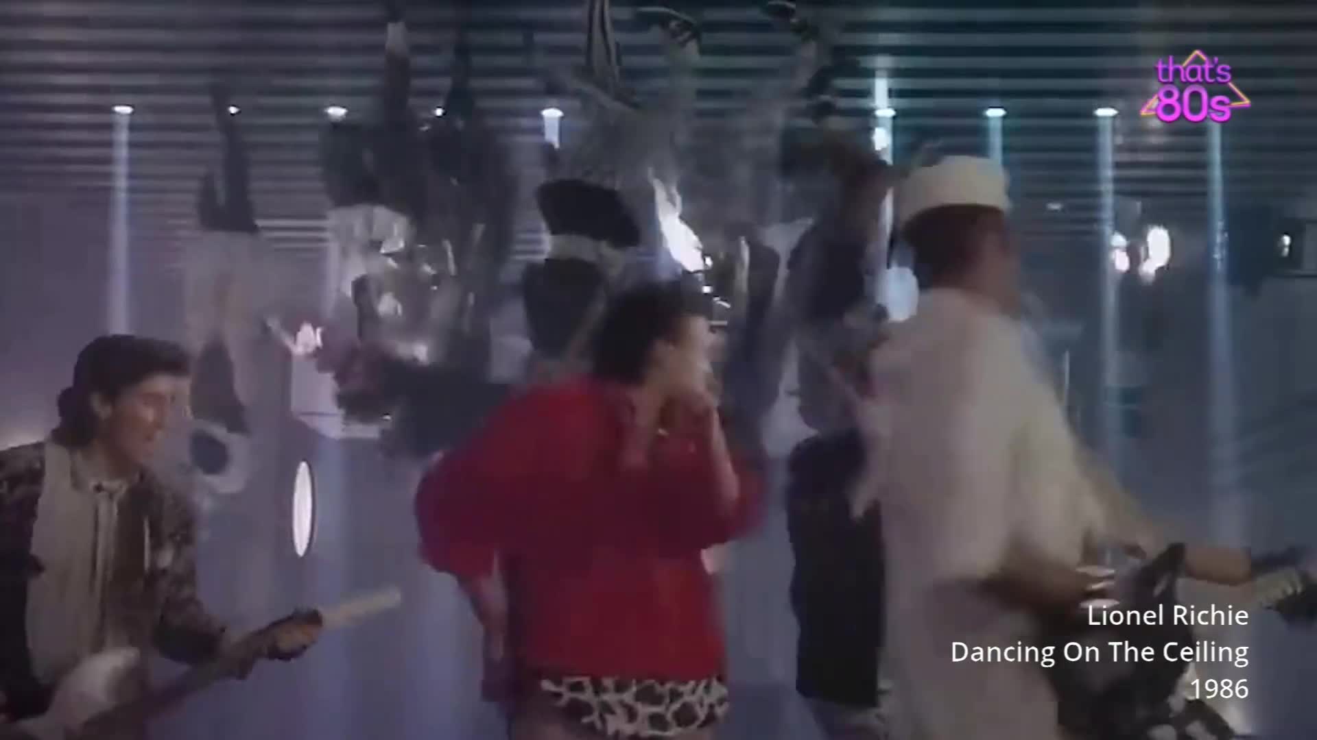 A group of people are dancing in a room with a striped ceiling. One man in a white outfit and cap is playing a guitar.