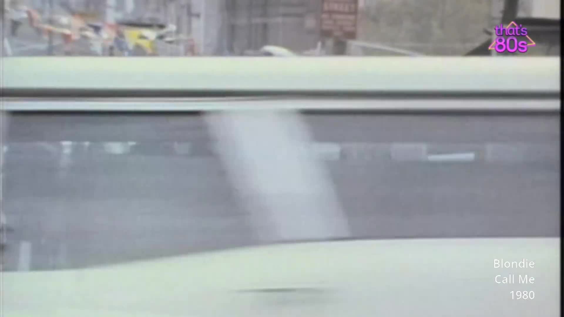 A pale yellow car drives past, its windows reflecting the grey street. A hint of bunting hangs in the background, suggesting a street party or event.