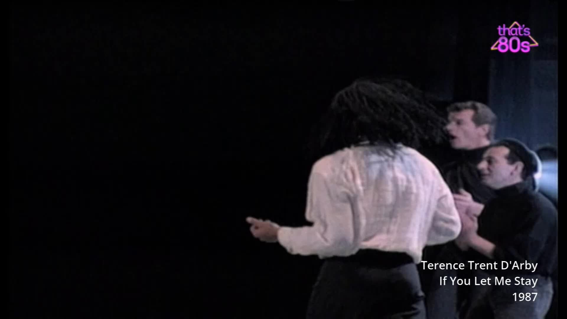 A woman with dark, bushy hair turns her back to me, her white shirt catching the light. Two men stand behind her, one with his mouth open as if singing.