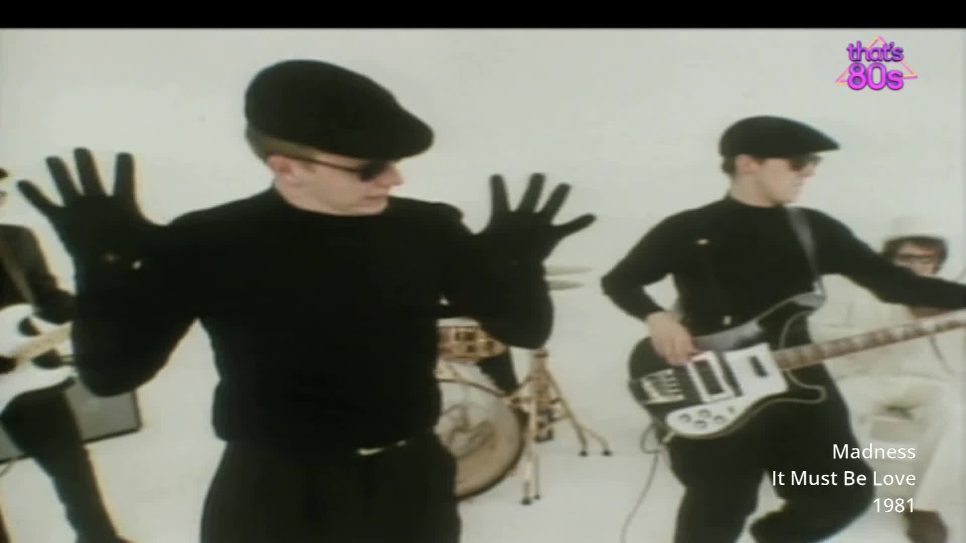 A band in black, complete with flat caps and dark gloves, performs on a stark white stage. The bassist, wearing sunglasses, strums his guitar with a focused intensity.