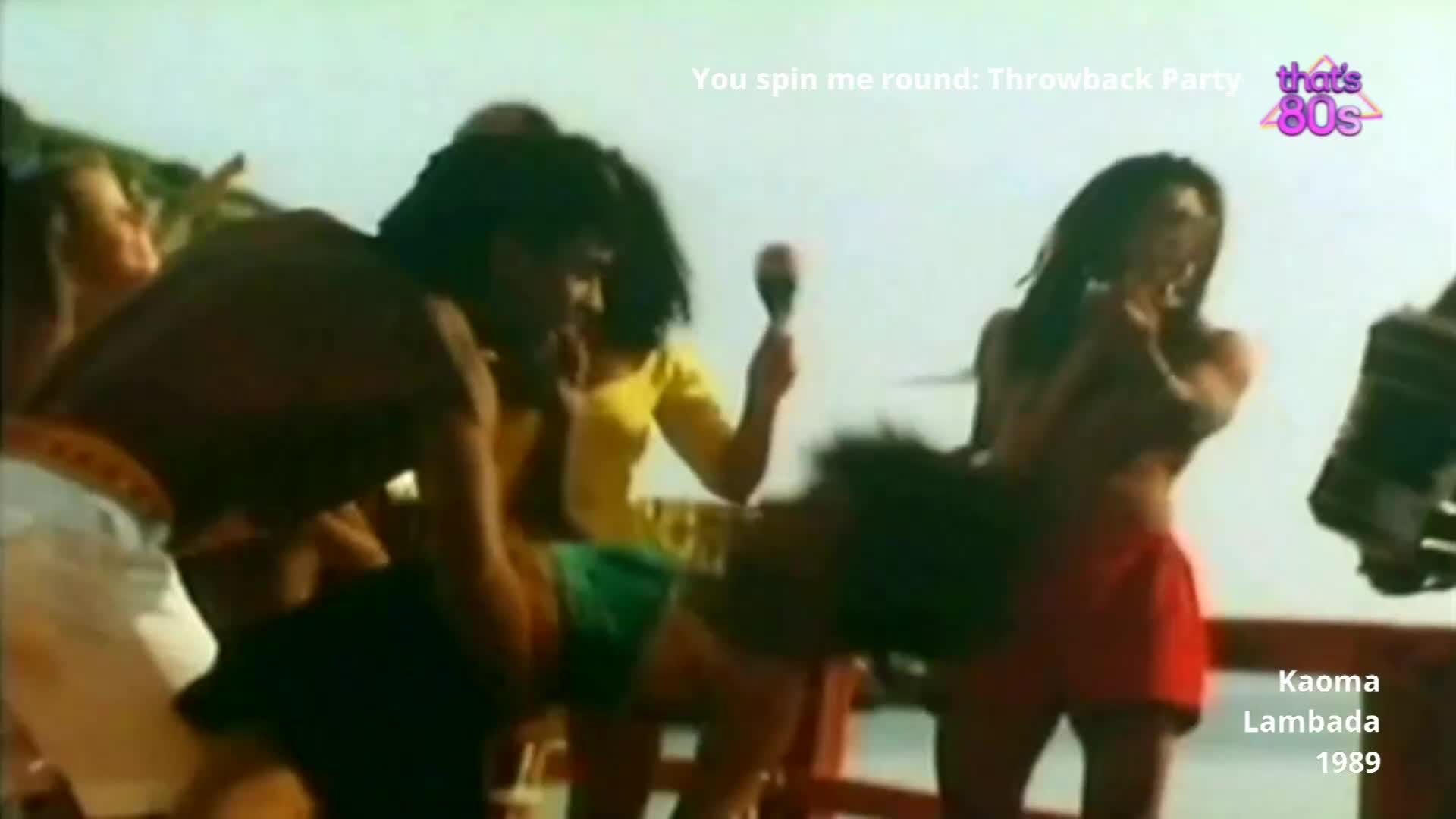 A group of people are dancing on a deck, their movements energetic. One woman in a yellow top shakes a maraca, while another in a red skirt spins.