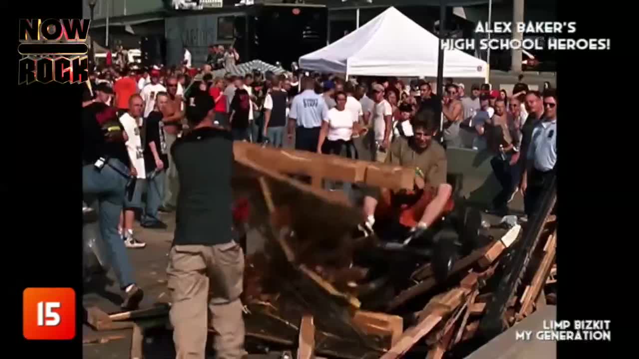 A man is using a chainsaw to cut through a pile of wooden planks, sending splinters flying. The crowd in the background seems to be watching a performance, judging by the tent and the stage setup.
