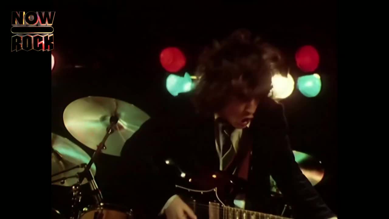 The guitarist, dressed in a dark suit, is hunched over his instrument, hair flying. His fingers dance across the fretboard as he belts out a song under the stage lights.

