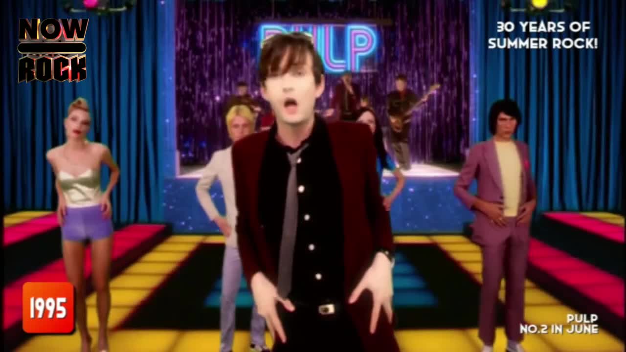 Jarvis Cocker, in a burgundy jacket, is singing with his arms out, while the band and some dancers stand behind him. The stage is lit up with bright colours and a backdrop of blue curtains.
