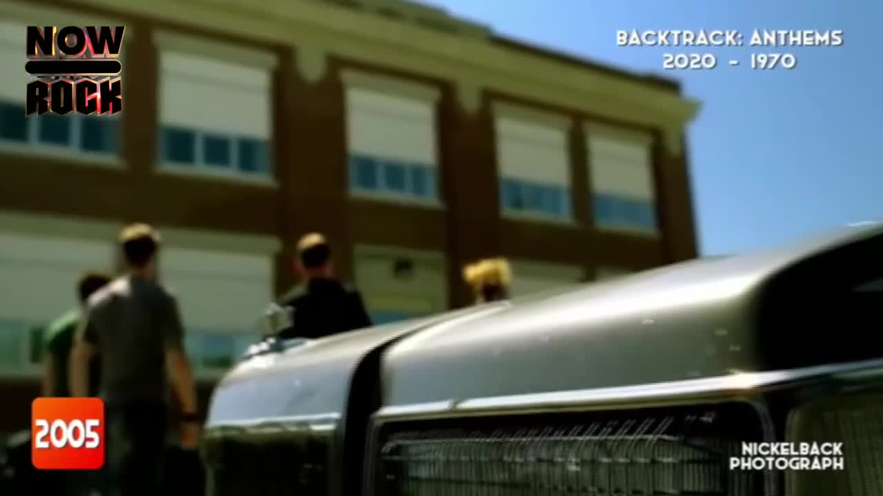 A group of guys are walking towards a classic car, possibly a shoot for a NOW Rock video. The camera focuses on the car's hood, with the school in the background.
