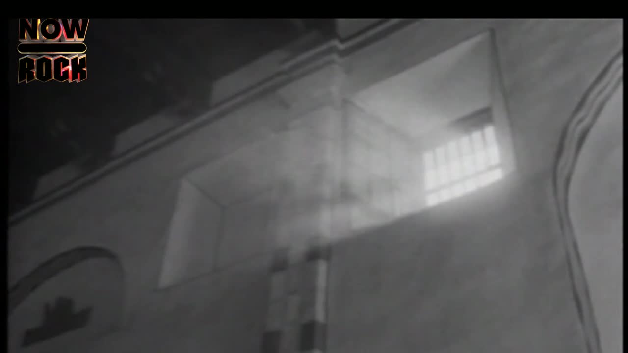Light streams through a barred window, illuminating a hazy interior. The stark, geometric lines of the building's architecture are softened by the diffused light.
