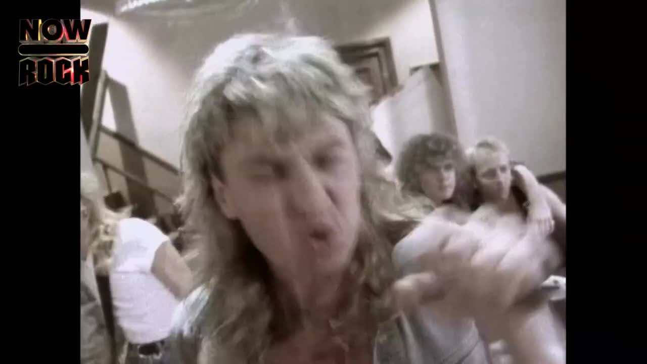 The lead singer, with his long, blonde hair, is right in front of me, singing into the camera with a lot of energy. Behind him, a couple of people are leaning in, watching from the side.
