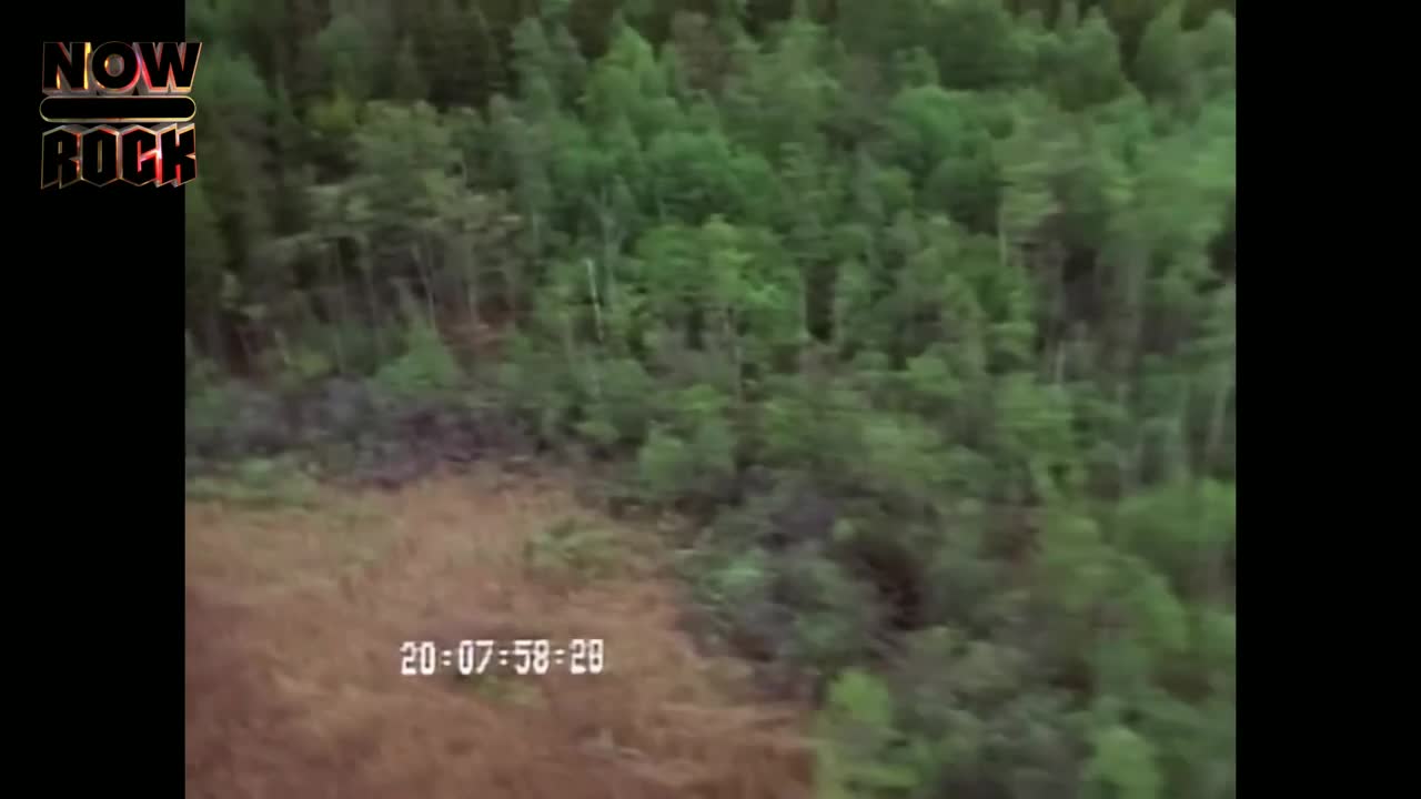 Dense green trees and a patch of dry, brown land fill the screen. The NOW Rock logo sits in the top left corner, and the time code is displayed near the bottom.
