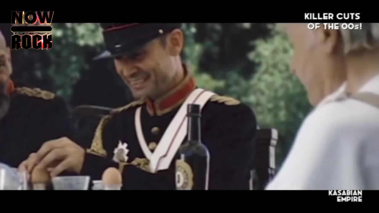 A man in a military uniform grins, reaching for a boiled egg on the table. The scene is set outdoors, with a bottle of something dark and a few other people at the table.

