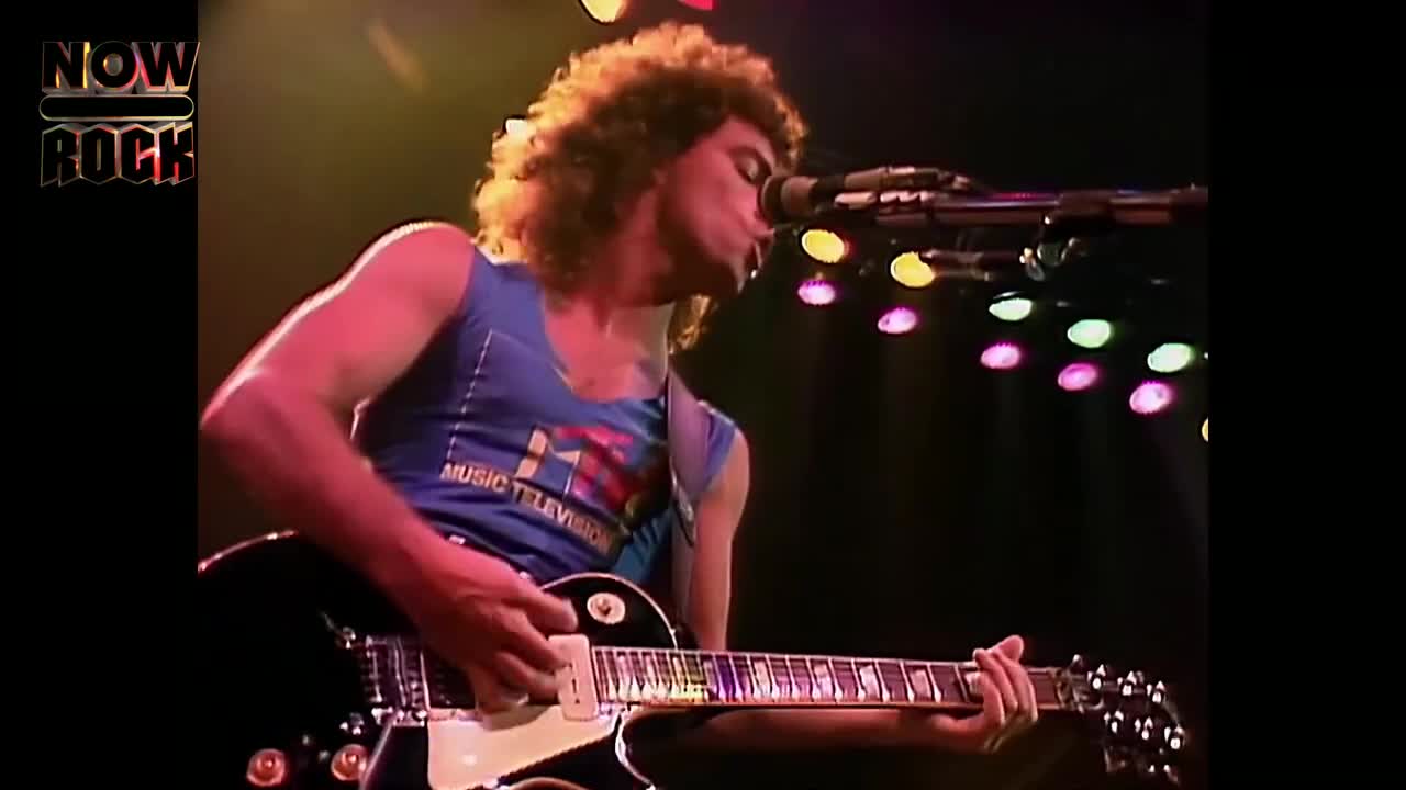 A guitarist, clad in a blue tank top, is singing into a microphone while strumming a black electric guitar. Above, the stage lights shine down, illuminating the singer's curly hair.
