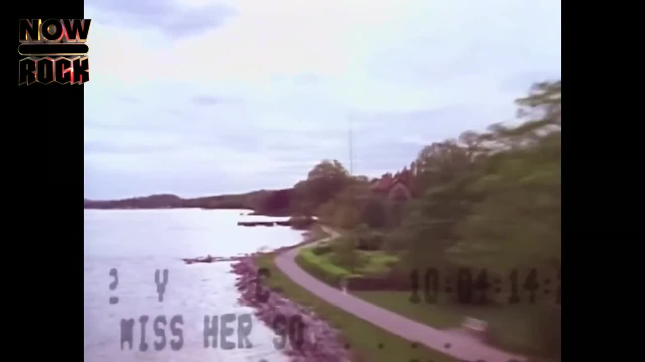The camera glides along the shoreline, capturing a calm lake and a winding path.  Text flashes on the screen, obscuring the view of the water.
