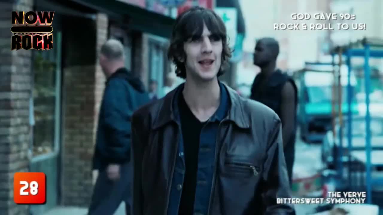 A man in a leather jacket walks toward the camera, his mouth open as if singing. Passersby move along the sidewalk behind him, the buildings of a UK street providing the backdrop.
