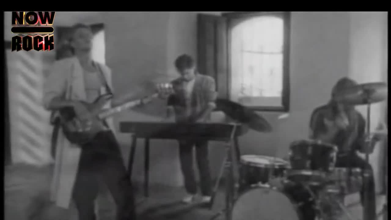 A band is playing, the bassist in a long coat strums his instrument, while the keyboardist and drummer keep the rhythm. The music is lively, and the scene is bathed in natural light from a window.
