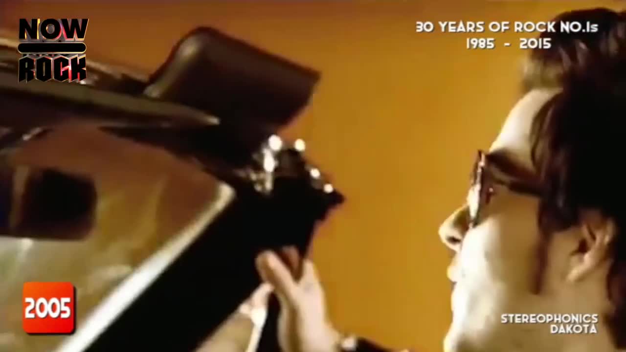 A man in glasses is reaching up, his hands visible as he interacts with the roof of a car. The "NOW Rock" logo and the year 2005 are visible on the screen.
