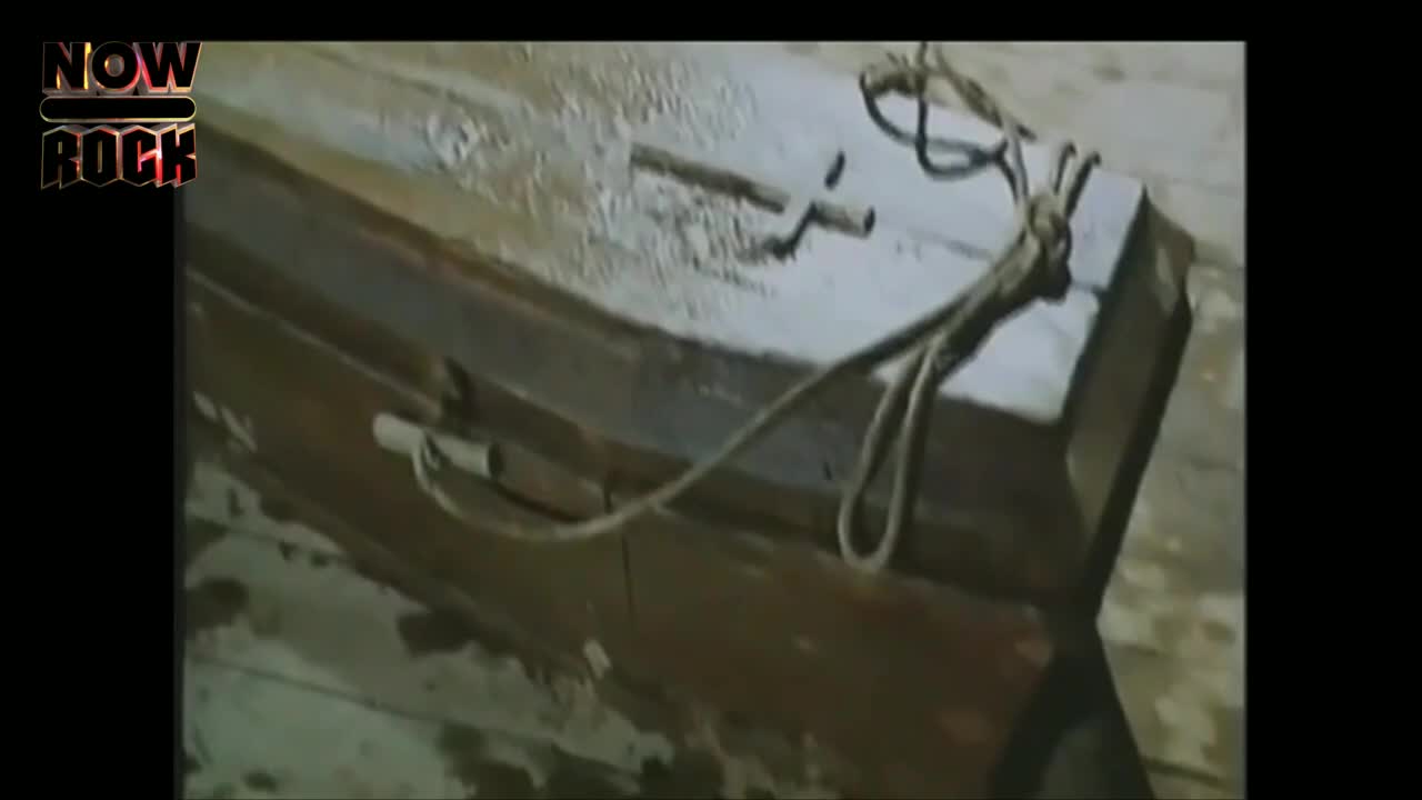 A wooden coffin lies on a surface, its lid secured by a rope. A small cross adorns the top, and the whole thing looks weathered.
