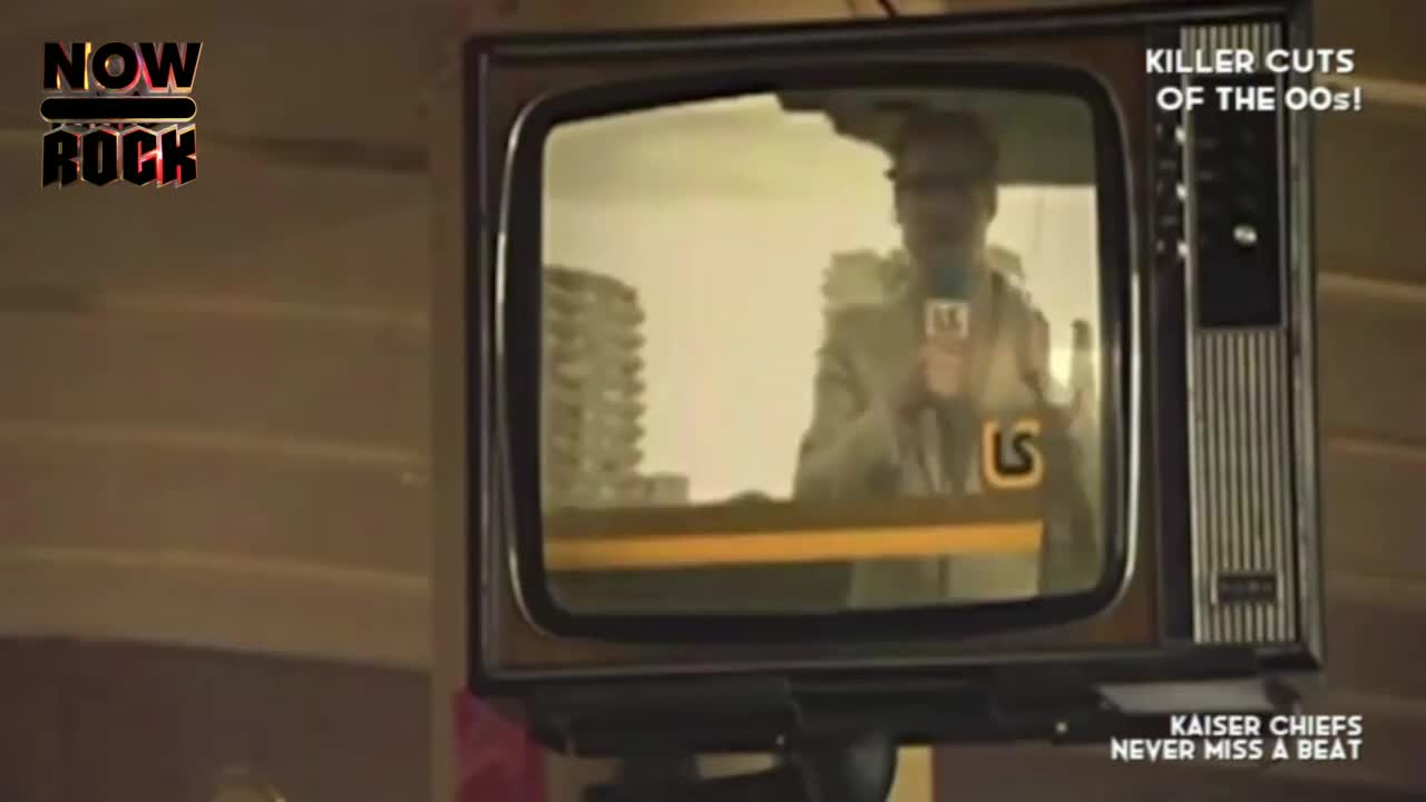 A vintage television sits on a wooden surface, displaying a man holding a microphone. The screen is showing what looks like a news report, with a building in the background.
