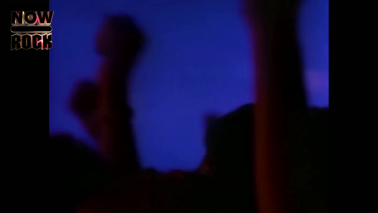Silhouetted figures are raising their arms, bathed in a deep blue light. The stage is dark, but the energy is palpable.
