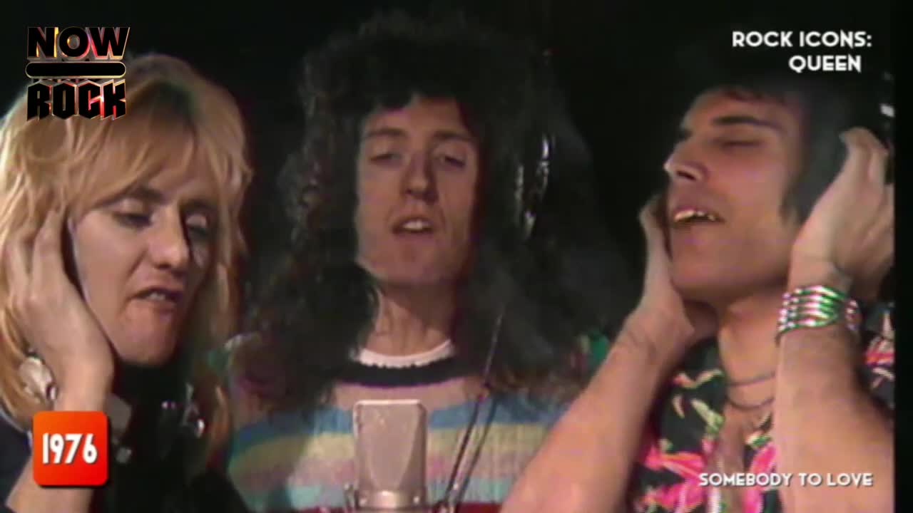Three members of Queen, all with headphones on, are singing into a microphone. The one in the middle, with long, curly hair, is looking up, while the others are gazing off to the side.
