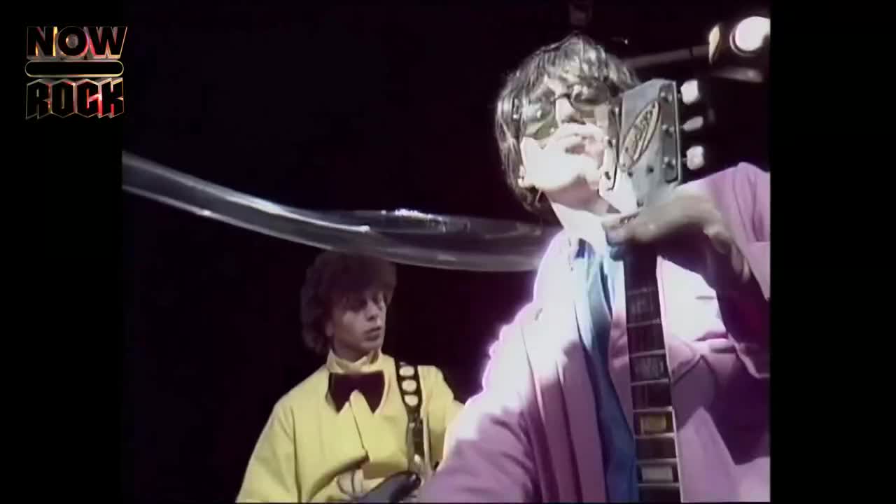 The guitarist, in a pink jacket and shades, holds his instrument close to his face, looking up. A second musician, in a yellow shirt, is on his right, ready to play.
