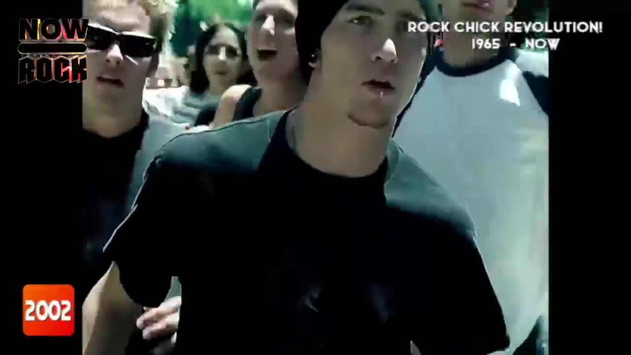 A group of young people are moving forward, the camera focused on a man with a lip piercing. The "NOW Rock" logo and the year 2002 are visible in the corner.
