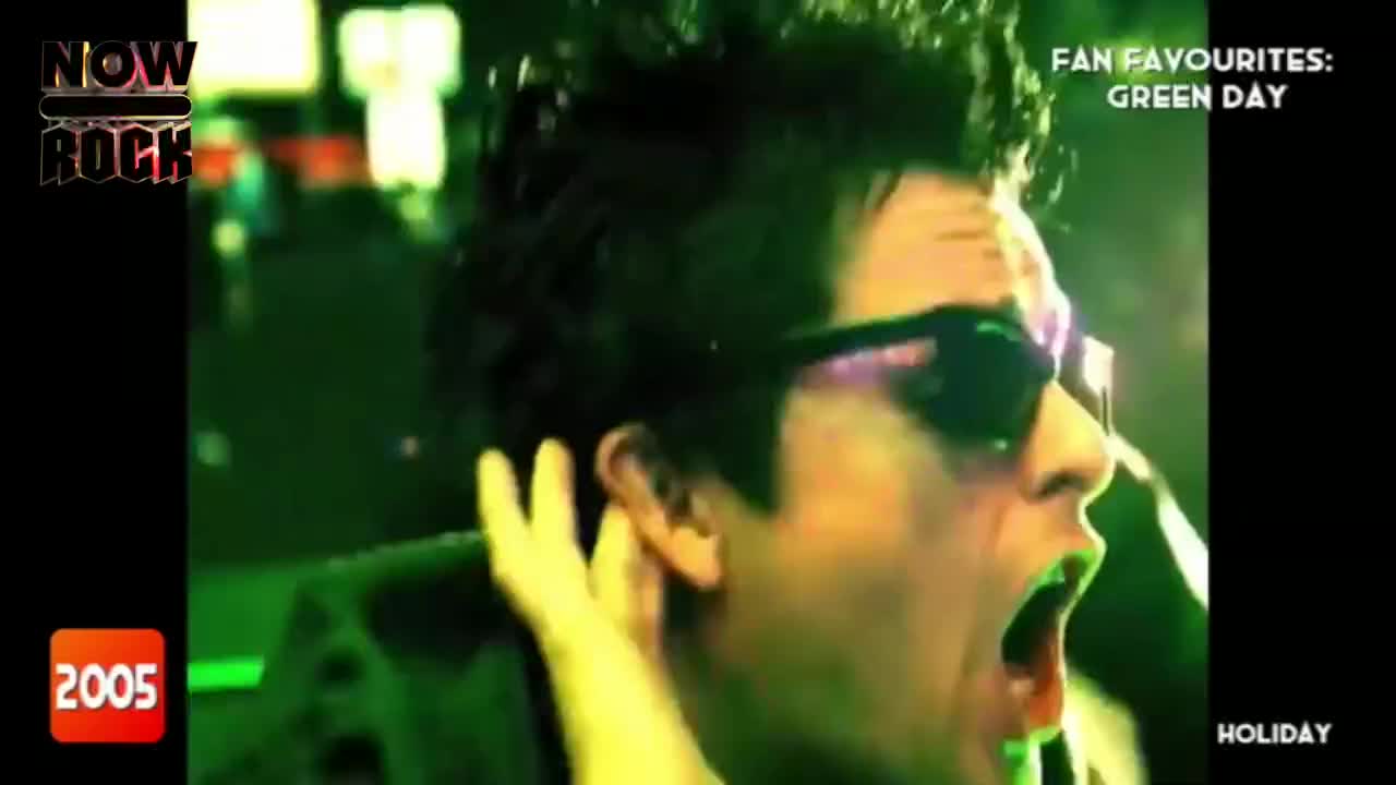 The singer, in dark sunglasses, is mid-scream, mouth wide open. The green-tinted lights of the NOW Rock broadcast illuminate his face.
