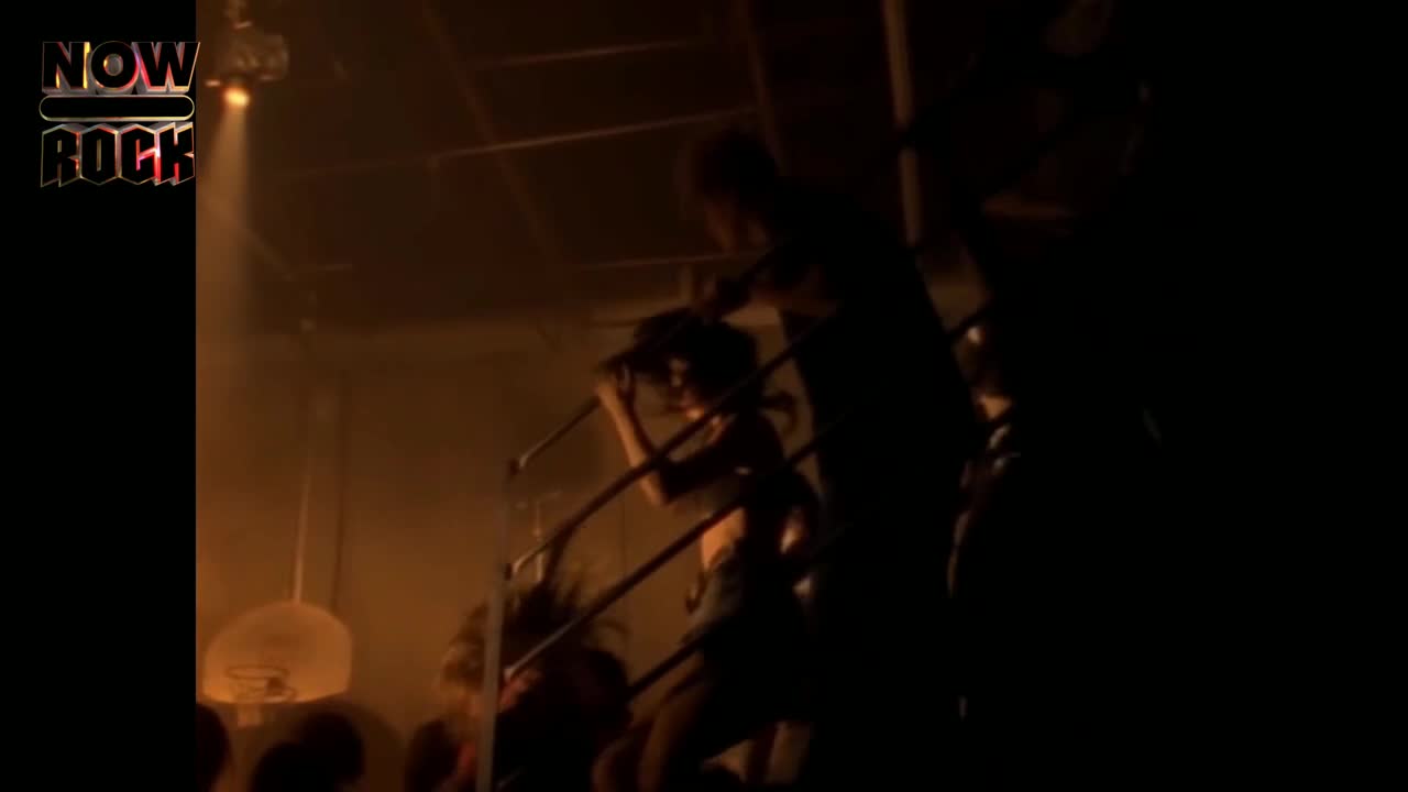 A figure leans against the railing, singing into a microphone. The stage is dim, but the light catches the singer's hair as they move.
