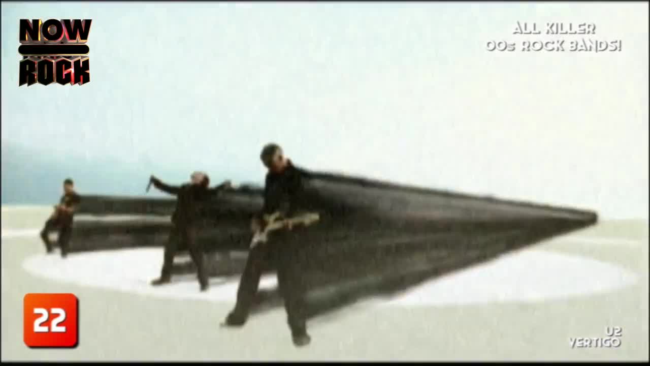 The band is playing on a stark, white stage, with a giant, dark shape behind them. The guitarist in the foreground is leaning into his instrument, while the singer gestures with a microphone.
