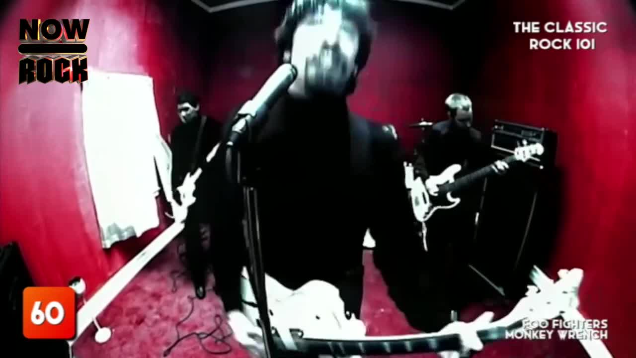 The lead singer, in a black turtleneck, is belting into a microphone, his guitar held low. Two other musicians, also in black, stand behind him, their instruments ready.
