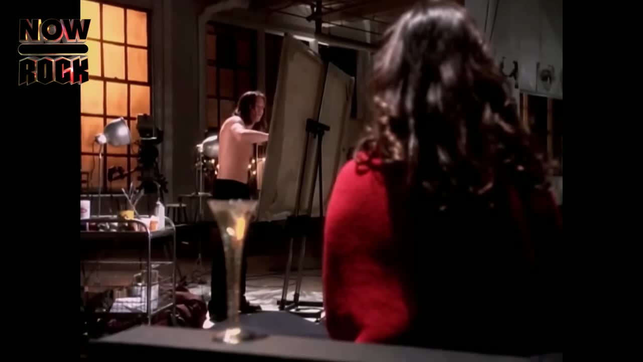 A shirtless man, brush in hand, works on a canvas; the scene is lit by warm light from the window. A woman in a red top is turned away, watching him.
